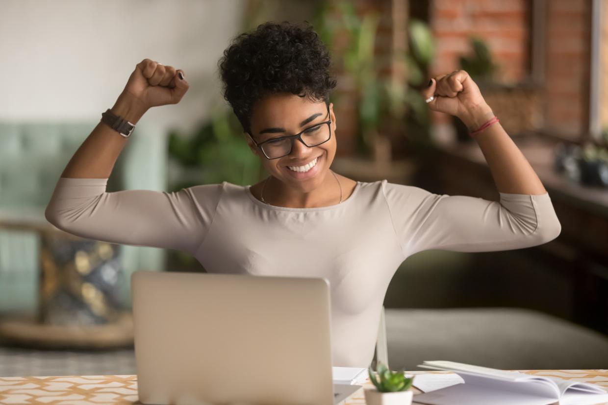 Young woman wearing glases sitting at laptop smiling with arms up in the air triumphantly